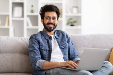 Uzak bir iş. Mutlu Hintli serbest çalışan adam evde dizüstü bilgisayar kullanıyor, klavyede daktilo kullanıyor, genç doğu çocuğu oturma odasında oturuyor, uzaktan kumandayla çevrimiçi proje üzerinde çalışıyor, boşluğu kopyalıyor.