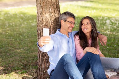 Romantik olgun çift parkta dinlenirken akıllı telefondan selfie çekiyor, mutlu orta yaşlı koca ve eş dışarıda ağaç altında dinleniyor, cep telefonuyla fotoğraf çekiyor ve sarılıyorlar.