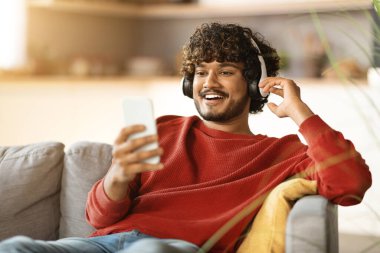 Harika bir uygulama. Mutlu Hintli adam evde dinlenirken akıllı telefon kullanıyor, cep telefonlu neşeli doğu çocuğu müzik akışı uygulaması, favori şarkıların tadını çıkarıyor, kopyala Space