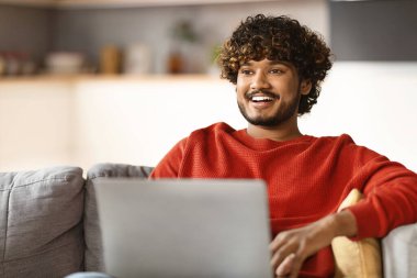 Bağımsız Kavram. Evde dizüstü bilgisayarlı mutlu doğu çocuğu koltuğunda oturuyor, oturma odasında koltukta oturmuş uzaktan kumandayla eğlenirken Hintli serbest çalışan genç adam gülümsüyor.