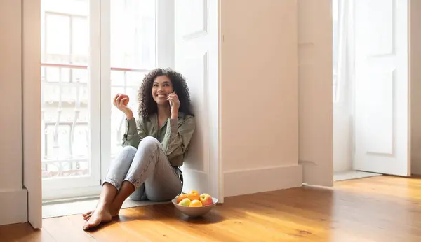 幸せなアフリカ系アメリカ人の女性チャット 携帯電話のフルーツを保持する 近代的なリビングルームで床の近くの窓かバルコニーのドアに座る モバイルオファー広告のための無料スペース — ストック写真