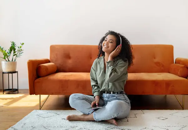 アフリカ系アメリカ人女性 デジタルタブレットでリラックス コンピュータオンラインでお気に入りの音楽を聴く ソファーの近くに座って ホームで週末の時間を過ごす — ストック写真