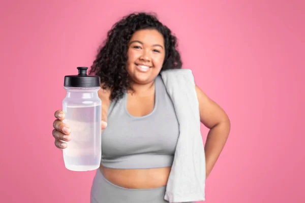 Cheerful black plus size lady in sportswear with towel showing bottle of water and smiling, resting after workout isolated on pink studio background. Recommendation for workout, fitness, weight loss