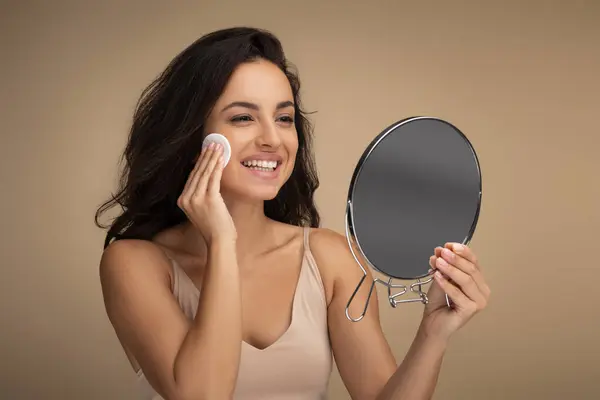 Smiling pretty young brunette indian woman holding small mirror and using cotton pads, hydrating face skin with toner, erasing makeup with micellar water, beige studio background, copy space, closeup