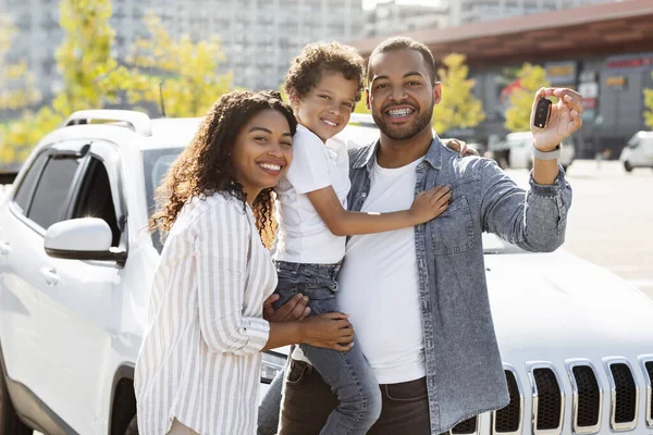 Happy african american family posing outdoors next to their brand new automobile, showing key. Cheerful loving black father, mother and son standing by nice white auto on parking spot. Family car