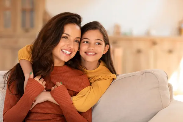 Loving Family. Portrait Of Young Mommy And Her Preadolescent Daughter Hugging Looking Aside, Posing Together Cuddling On Sofa Indoor. Kid Girl Embracing Mom From Back. At Home