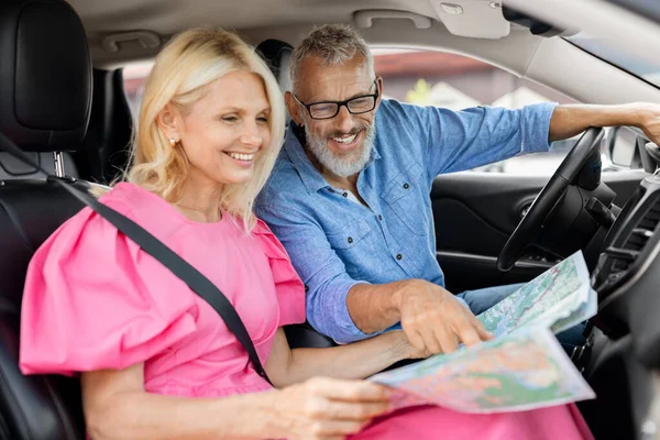 Car ride, travelling, tourism concept. Excited cheerful smiling attractive retired man and woman travelers sitting inside auto, reading map, senior couple choosing destination to travel