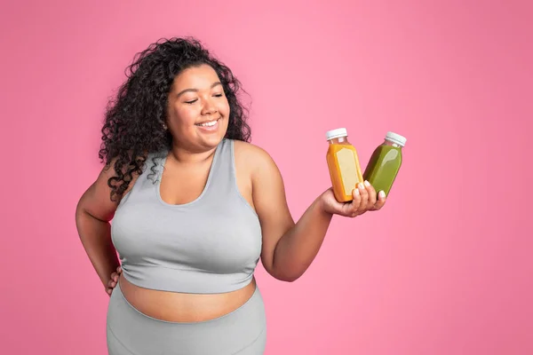 Happy black body positive lady in sportswear holding and looking at bottles of juice, fresh during workout, isolated on pink studio background. Advice of proper nutrition, weight loss and diet