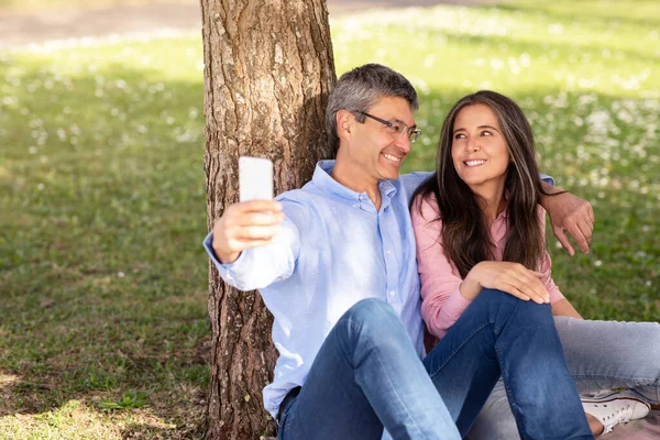 Romantic Mature Couple Taking Selfie On Smartphone While Relaxing In Park Together, Happy Middle Aged Husband And Wife Resting Under Tree Outdoors, Embracing And Capturing Pictures On Mobile Phone