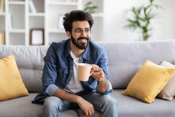 英俊潇洒的印度人 一杯咖啡放松在家里的沙发上 年轻的东方人 戴着眼镜 拿着杯热饮 远眺远方 享受家庭休闲 复印空间 — 图库照片