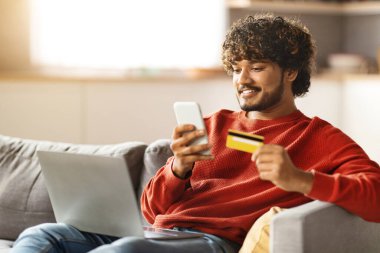 Mobil Banka Uygulaması. Evde koltukta otururken telefon ve kredi kartı kullanan gülümseyen Hintli adam internetten alışveriş yapıyor, akıllı telefon için modern uygulamalara bakıyor, boş alan arıyor.