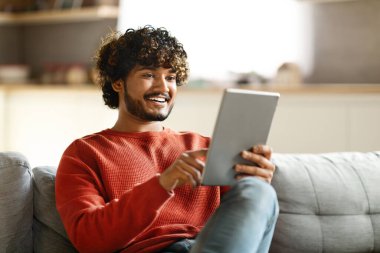 Dijital tabletli yakışıklı genç Hintli adam evdeki kanepede dinleniyor, modern aletlerle oturma odasında dinleniyor, internetten alışveriş yapıyor ya da internette geziniyor, boşluğu kopyalıyor.