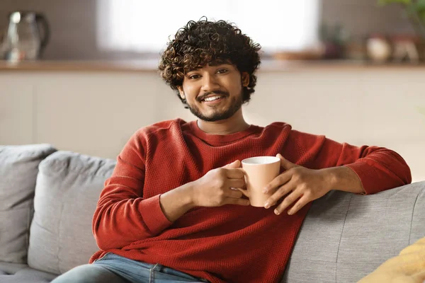 快乐的印度年轻人 喝着一杯茶 舒服地躺在沙发上 英俊的千禧年东方人 拿着杯子 在相机前微笑 享受家庭休闲 复印空间 — 图库照片