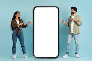 Güzel online teklif. Bin yıllık olumlu Hintli çift stil sahibi erkek ve kadın reklam için beyaz boş ekran kopyalama alanı ve mavi arka plan ile büyük telefona bakıyor.
