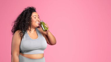 Spor kıyafetleri içinde bir şişe meyve suyu ya da smoothie içen, pembe stüdyo arka planında izole edilmiş poz veren şişman bir kadın, panorama, boş alan. Kilo kaybı, vücut bakımı, reklam ve teklif, vitaminler.