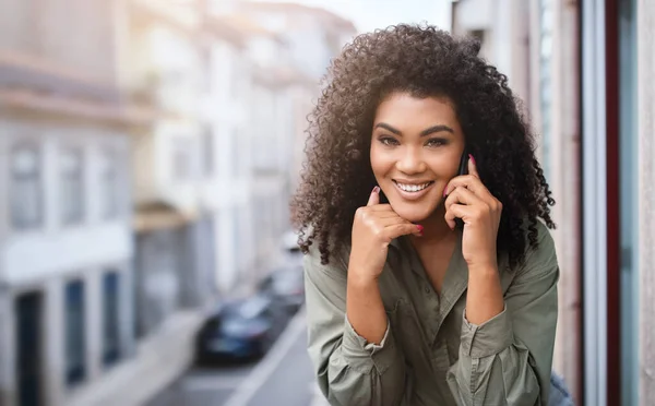 Great mobile connection. Happy young african american woman enjoying phone talk with friend via smartphone, smiling to camera posing at balcony outside. Empty space for text