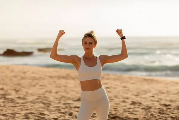 Positive strong european woman in sportswear doing success and victory gesture, showing muscle biceps on ocean beach during training. Morning workout, body care outdoors