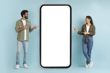 Güzel cep telefonu teklifi. Mutlu çok ırklı çift, beyaz ekranlı büyük cep telefonunu işaret ederek, telefon uygulaması ya da web sitesi tasarımı için boş alan gösteriyor, mavi arkaplan, kopyalama alanı