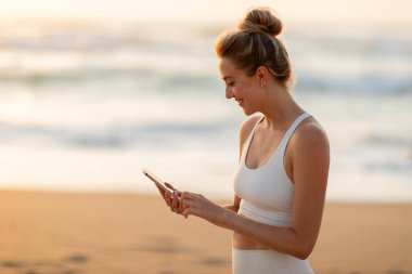 Spor giyimli neşeli Avrupalı bayan sporcu, cep telefonu kullanıyor, açık havada idmandan sonra deniz sahilinde duruyor. Spor, vücut bakımı uygulaması, sosyal ağlar, blog