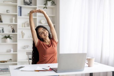 Masasında dizüstü bilgisayarı ve kırtasiyesiyle oturan neşeli, çekici, genç Hintli kadın ev ofisinde vücudu esnetiyordu. Serbest çalışan, çalışmalarının sonuçlarından memnun hissediyordu.