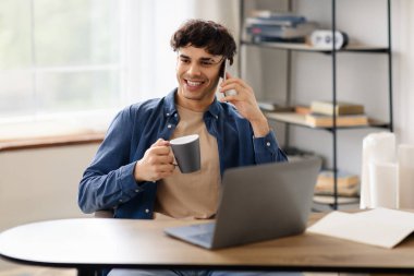 Gülümseyen Orta Doğulu Genç İşadamı Laptop 'ta gezinirken telefonda konuşuyor, kahve içiyor, Modern Ofis Masasında Başarılı Çalışma Gününün tadını çıkarıyor. İş İletişimi