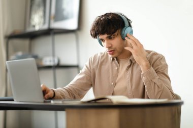 Orta Doğulu genç adam, kablosuz kulaklık takıyor ve dizüstü bilgisayarında internete bakıyor, modern ofiste masada oturuyor, çevrimiçi çalışırken müzik dinliyor ve çalışıyor.