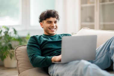 Neşeli Arap milenyum adamı dizüstü bilgisayarda yazıp çalışıyor oturma odasında uzaktan çalışma tarzının tadını çıkarıyor bilgisayarla çalışıyor kanepeye uzanıyor ve gülümsüyor. Serbest meslek.