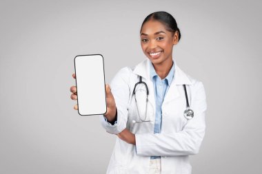 Beyaz önlüklü neşeli siyahi kadın doktor, ekranı boş, gri arka plan stüdyosuna izole edilmiş, pozitifliğini ifade eden akıllı telefonu tutuyor. Sağlık hizmetleri, reklam ve teklif, uygulama tavsiyesi