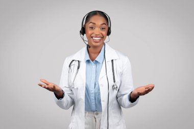 Resimde gülümseyen profesyonel siyahi kadın doktor ellerinde boş bir alan, gri arka planda, stüdyoda izole edilmiş. Uzaktan tıbbi yardım, modern sağlık hizmetleri, klinikte destek
