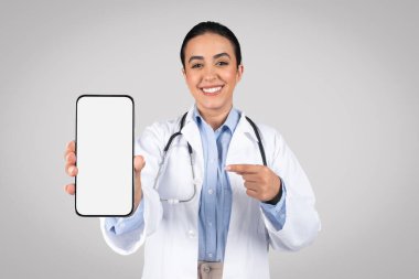 Latin kadın doktor cep telefonuna boş ekranla işaret ediyor, promosyon tıbbi teklifleri veya mesajlar için kişiselleştirilebilir alan sunuyor, gri stüdyo arka planına karşı.