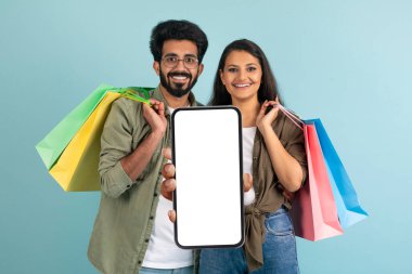 Neşeli genç Hintli kadın ve erkek alışverişler yapıyor, beyaz ekranlı ve gülümseyen akıllı telefonları gösteriyor, güzel alışveriş mobil uygulaması kullanıyor, mavi arka plan, kopyalama alanı kullanıyor.