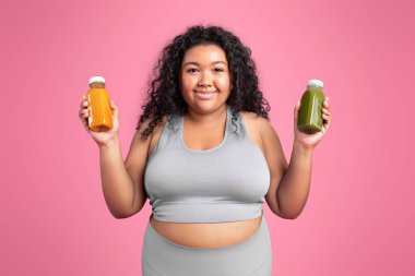 Spor giyim bölümünde iki şişe smoothie gösteren, pembe stüdyo arka planında kameraya poz veren ve gülümseyen büyük beden bir kadın var. Seçenek ve diyet, kilo kaybı, reklam ve teklif