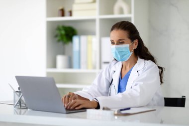 Tıbbi üniforma giyen ve maske takan olgun bir kadın doktor masasında oturuyor, bilgisayarında çalışıyor, uzayı kopyalıyor. Soğuk algınlığı döneminde doktorların hastalara danışması