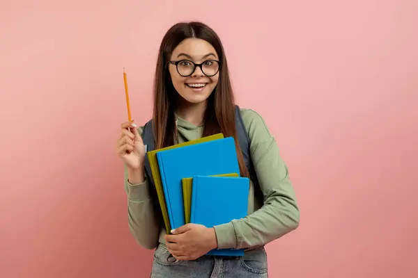 带着背包和练习本的快乐的少年学生有一个聪明的想法 快乐美丽的少年女子举起铅笔 经受着灵感的时刻 在工作室里摆出粉红的背景 抄袭空间 — 图库照片