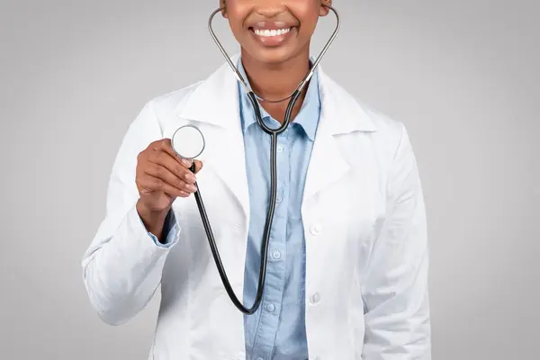 Happy black woman doctor listening through stethoscope, attentive healthcare, expertise in diagnosis, approach to patient, medical examinations, isolated on gray background, studio, cropped