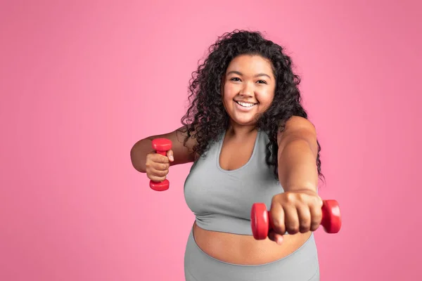 Cheerful excited black chubby lady in sportswear doing exercises with hand dumbbells, standing isolated on pink studio background. Workout, fitness, weight loss and pilates