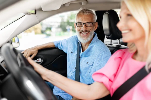 Cheerful senior man proud of his wife got driving license and drive car. Happy elderly husband looking woman driver and smiling, older couple going shopping together by auto