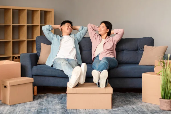 Real Estate Offer. Happy Asian Couple Takes a Restful Pause Leaning Against the Couch Moving New Rental Apartment, Relaxing Indoors Among Carton Boxes, Sharing a Moment of Happiness