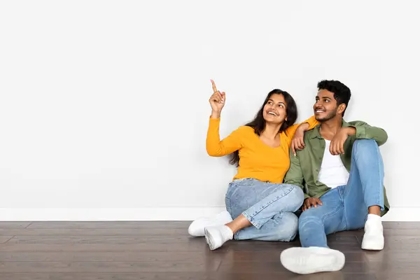 Happy young indian couple buying new house, sitting on floor in empty room in apartment, pointing at free space over white wall. Mortgage for millennials, rent concept