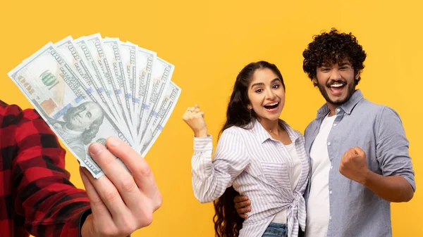 Happy indian couple celebrating success with raised fists, hand with many money, isolated on yellow background, studio. Win finance, savings, profit dollars, lottery and shopping