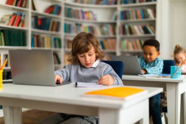 Akıllı okul çocukları, ilköğretim sınıfındaki masalarda oturuyorlar, dizüstü bilgisayarlarla eğitim içeriğini araştırıyorlar, dijital öğrenme dünyasını araştırıyorlar, eğitime çağdaş bir yaklaşımı işaret ediyorlar.