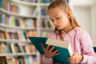 Odaklanmış liseli kız, kütüphanedeki bir kitaba hafifçe göz gezdiriyor. Huzurlu eğitim alanına dalmışken sayfalarda gezinen, meşgul, yansıtıcı bir öğrenme atmosferini somutlaştırıyor.