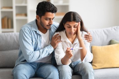 İstenmeyen hamilelik. Endişeli Hintli çift test sonuçlarına bakıyor. Stresli Hintli erkek ve kadın ebeveyn olmaya hazır değiller. Evdeki kanepede oturuyorlar.