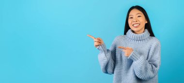 Mavi stüdyo arkaplanından fotokopi alanını gösteren kalın kazaklı mutlu genç Çinli kadın. Pozitif Asyalı kadın harika bir teklif sunuyor, reklam, panorama, web-pankart