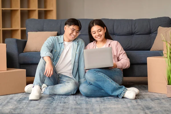 Asian Couple Using Laptop Websurfing And Searching New Rental Apartment, Using Online Service For Moving, Sitting Amid Packed Carton Boxes And Packages In Their Living Room At Home