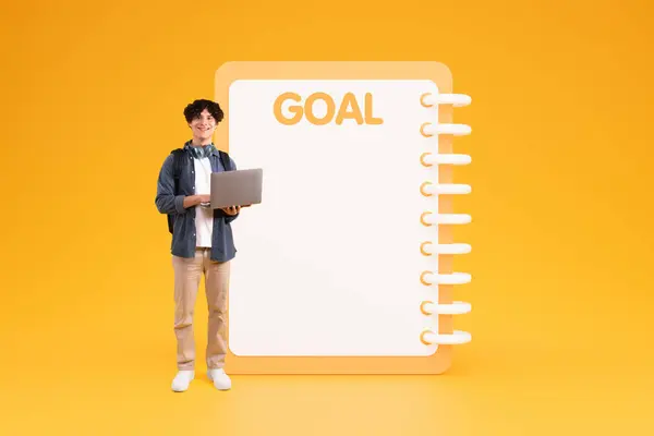 Smiling young caucasian guy student in casual use laptop for study near big list goal with free space, isolated on yellow studio background. Study, day planning, dream motivation