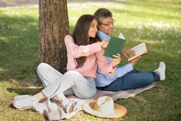 Happy loving middle aged couple sitting in park, reading books together, romantic mature man and woman relaxing on plaid under tree and quoting their favorite literature to each other