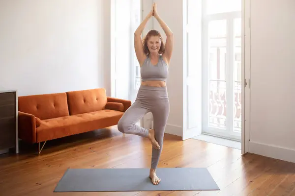 Smiling Senior Woman Standing In Tree Pose While Practicing Yoga At Home, Active Sporty Older Lady Exercising On Yoga Mat In Living Room, Enjoying Wellness And Healthy Lifestyle, Full Length