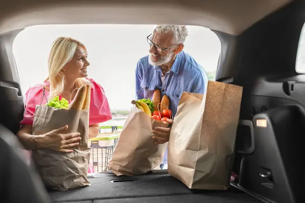 Loving attractive caucasian elderly couple putting food into car with open trunk, senior man and woman husband and wife load grocery packages in auto, shot from inside vehicle