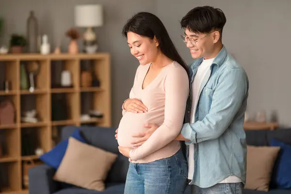 Pregnancy. Happy married japanese couple awaiting baby, standing together embracing in modern living room indoor, asian husband touching wifes belly. Family, joy of parenthood concept
